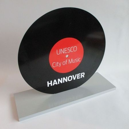 Eine stilisierte schwarze Schallplatte mit einem roten Etikett mit der Aufschrift "UNESCO City of Music Hannover" auf einem grauen Sockel, die Hannovers musikalisches Erbe symbolisiert.