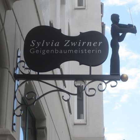 Schild für Sylvia Zwirner, Geigenbaumeisterin, mit der Silhouette einer Geigerin, die auf einem Gebäude vor blauem Himmel angebracht ist.
