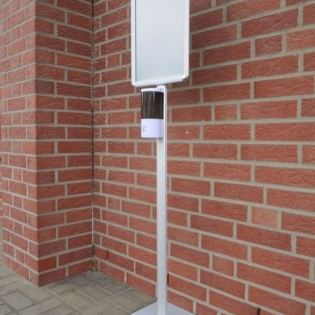 Weißer Informationsstand mit einer leeren Tafel und einem Desinfektionsmittelspender vor einer roten Backsteinwand.