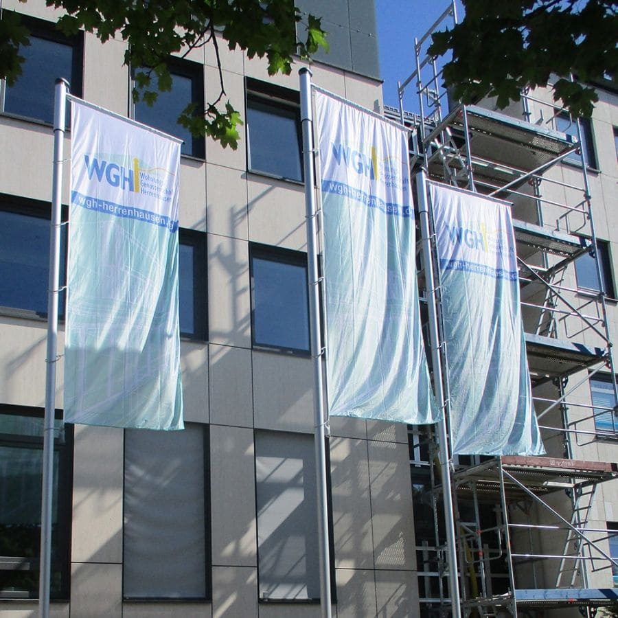 Drei große Banner mit dem WGH-Logo sind an Masten vor einem modernen Gebäude angebracht, im Hintergrund ist ein Baugerüst zu sehen.