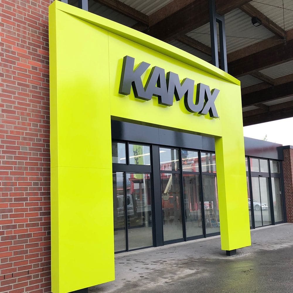 Leuchtend lindgrüne Schaufensterfront mit dem großen Schild "KAMUX" in modernen schwarzen Buchstaben über Glastüren. Im Hintergrund eine Backsteinmauer.